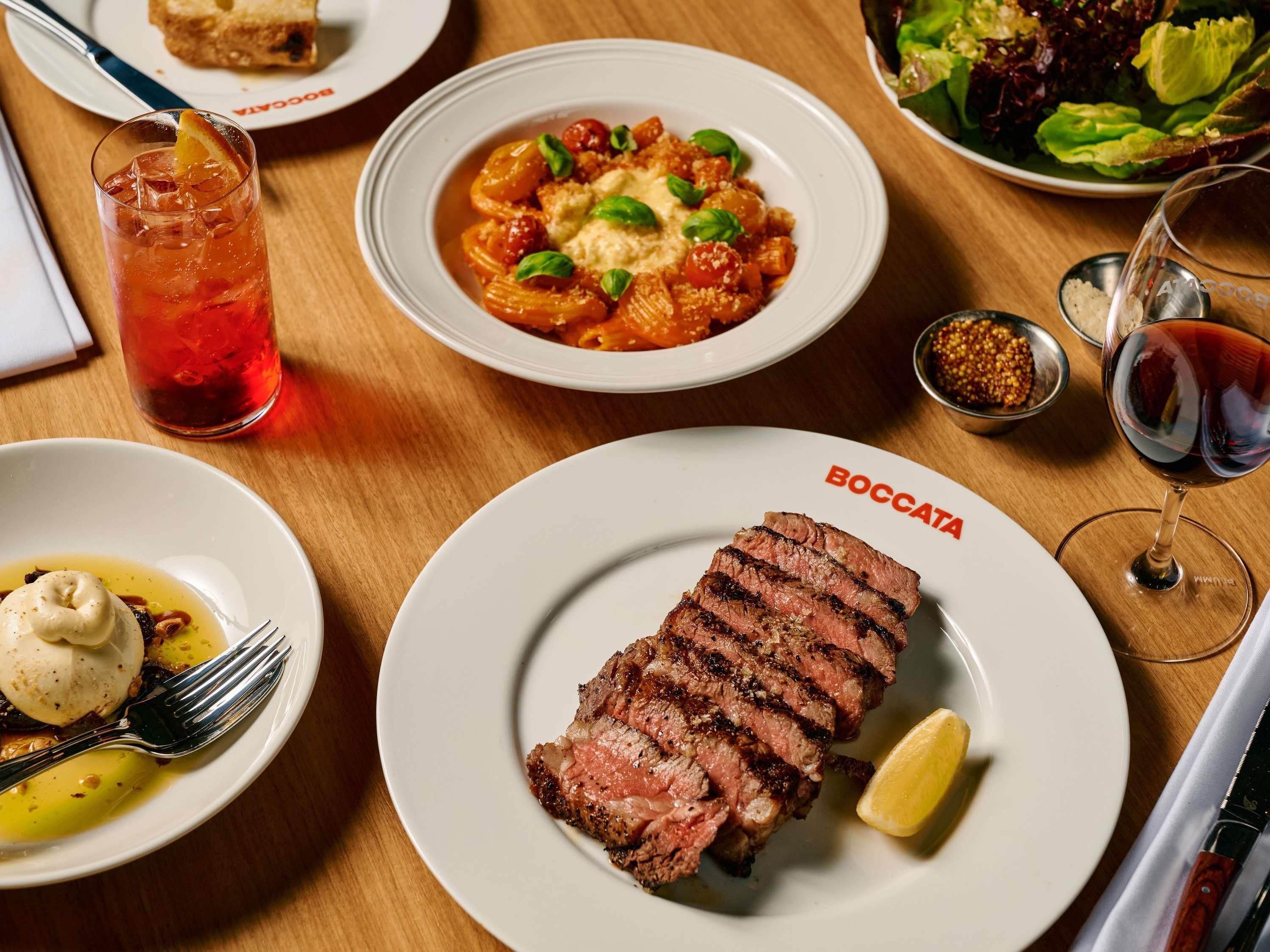 Een eettafel met een bord gesneden steak, een schaal pasta, een glas rode drank en een bord salade.