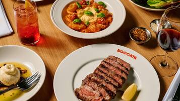 Una mesa de comedor con un plato de filete en rodajas, un bol de pasta, un vaso de bebida roja y un plato de ensalada.