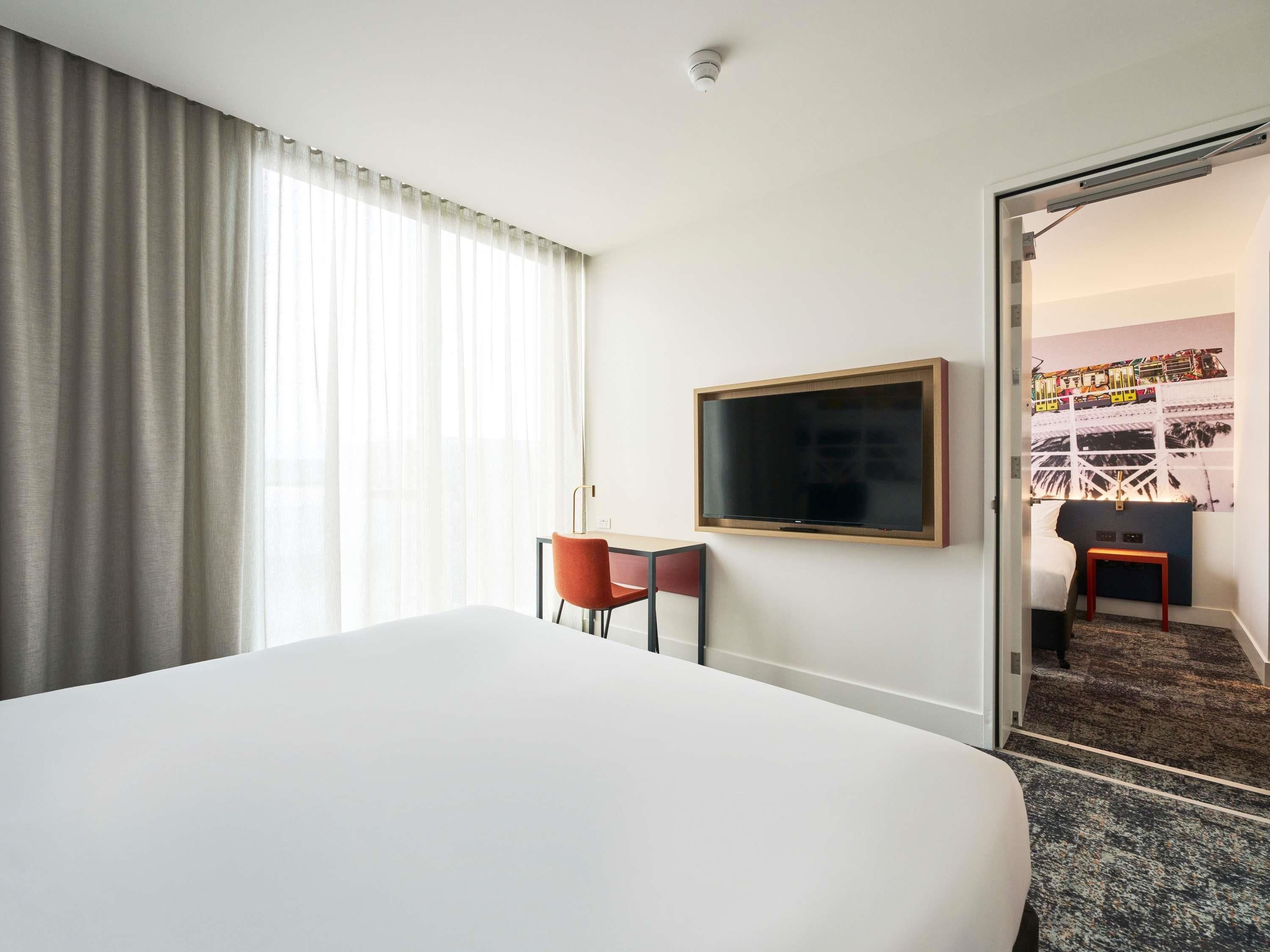 Une chambre d’hôtel avec un grand lit, un bureau avec une chaise, un téléviseur fixé au mur et une porte donnant sur une autre pièce.