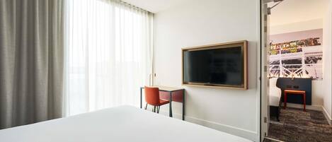 A hotel room with a large bed, a desk with a chair, a television mounted on the wall, and a door leading to another room.