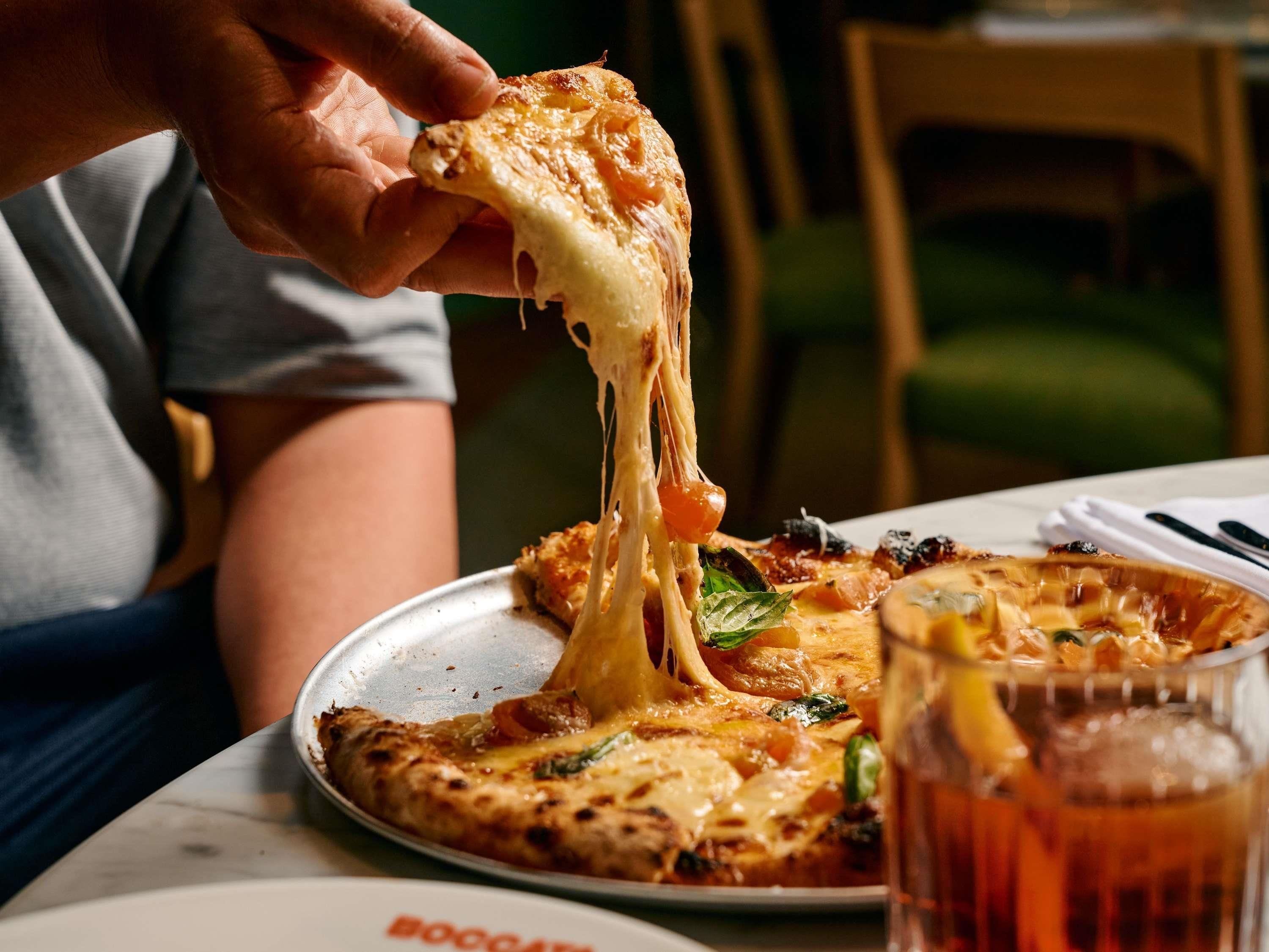 Una persona sostiene una porción de pizza con queso derretido, un vaso de bebida, y un plato con restos de pizza sobre una mesa.