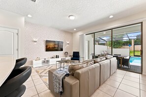 A modern living room with a sofa, a dining area, and a view of a swimming pool.
