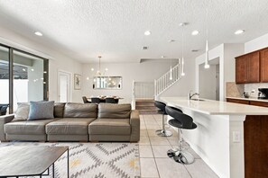 A modern living room with a sofa, a dining area, and a kitchen with a bar.