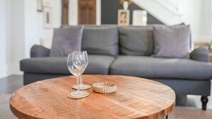Una mesa de centro redonda de madera con un tablero de vidrio transparente y dos posavasos tejidos, ubicada en una sala de estar con un sofá gris y una escalera al fondo.