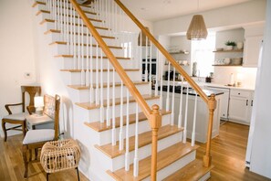 A staircase with wooden balusters and a handrail, leading to a kitchen area with white cabinets and a hanging pendant light. - Victorian Gem Steps from Main St - Sleeps 9 & Pets (Saint Charles)