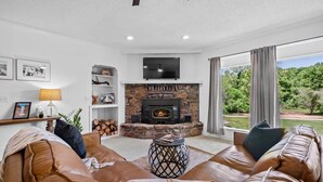 A living room with a stone fireplace, leather sofas, a TV mounted above the fireplace, and a view of greenery outside. - Creekside Oasis w/ Pool & Hot Tub near Beaver Lake (Rogers)
