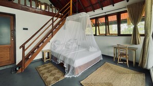 A room with a bed under a mosquito net, a wooden staircase, a small table, and a window with curtains.