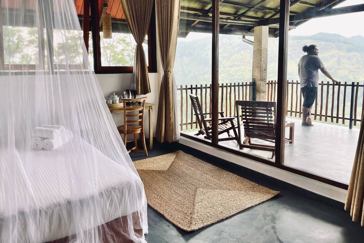 A room with a bed covered in white netting, a wooden table with a kettle, and a view of a mountainous landscape through large windows.