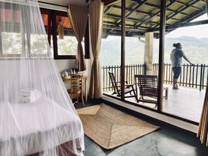 Une chambre avec un lit recouvert d’un ciel de lit blanc, une table en bois avec une bouilloire, et une vue sur un paysage montagneux par de grandes fenêtres.