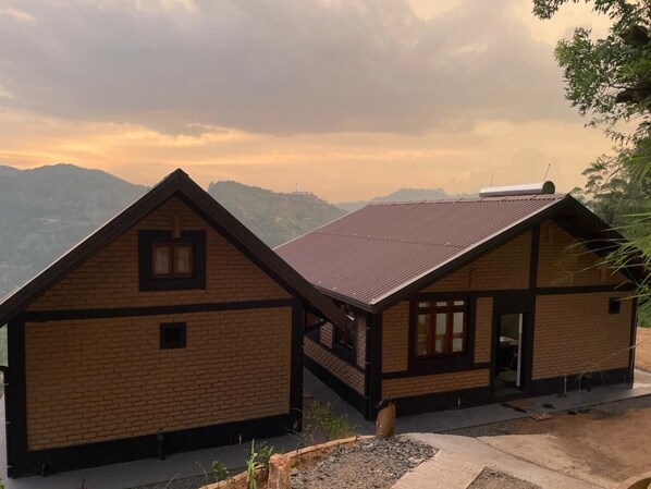 Une cabane en bois à deux étages, dotée d’un toit en pente, située sur une colline et offrant une vue sur les montagnes lointaines.
