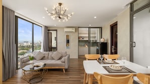 A modern living room with a sofa, dining table, and chairs. There is a kitchen area with cabinets and a refrigerator. The room has large windows and a chandelier. - Rose Sea Hotel & Apartment (Da Nang)