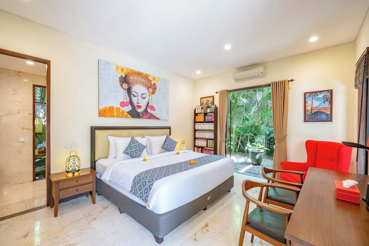 A hotel room with a large bed, a bedside table with a lamp, a bookshelf, a red chair, and a view of greenery through the window.