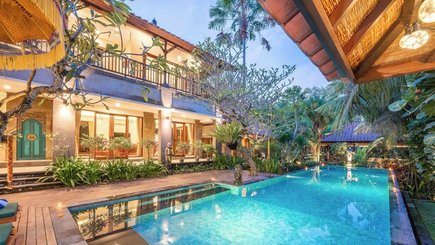Un área de piscina con cubierta de madera, un edificio de dos pisos con balcones y plantas tropicales.