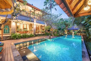 A pool area with a wooden deck, a two-story building with balconies, and tropical plants. - Puri Lana Villa by Nagisa Bali (Tabanan)