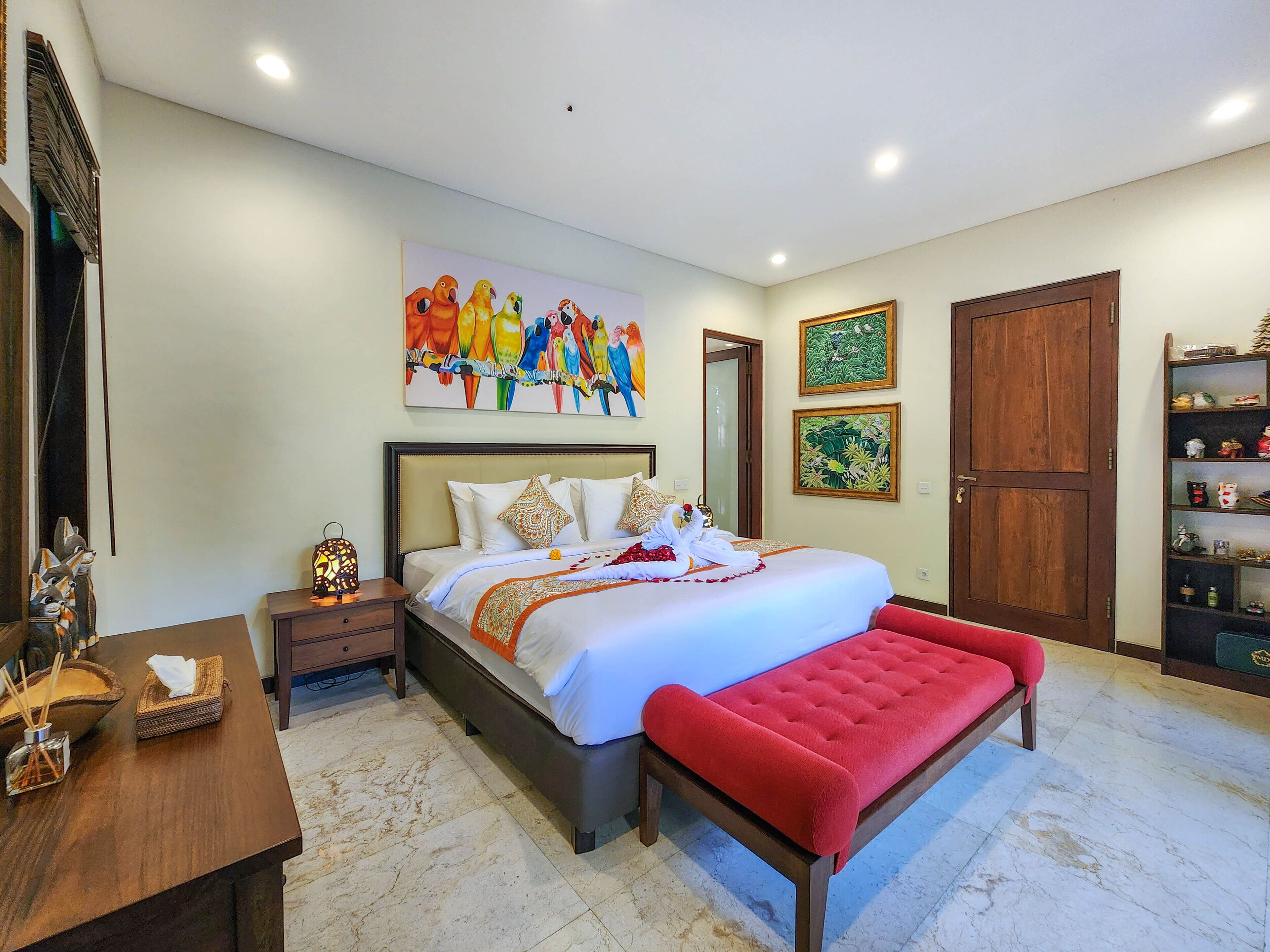 Un dormitorio bien iluminado con una cama grande, un banco rojo, una mesita de noche de madera y una pared con un cuadro de un guacamayo colorido.