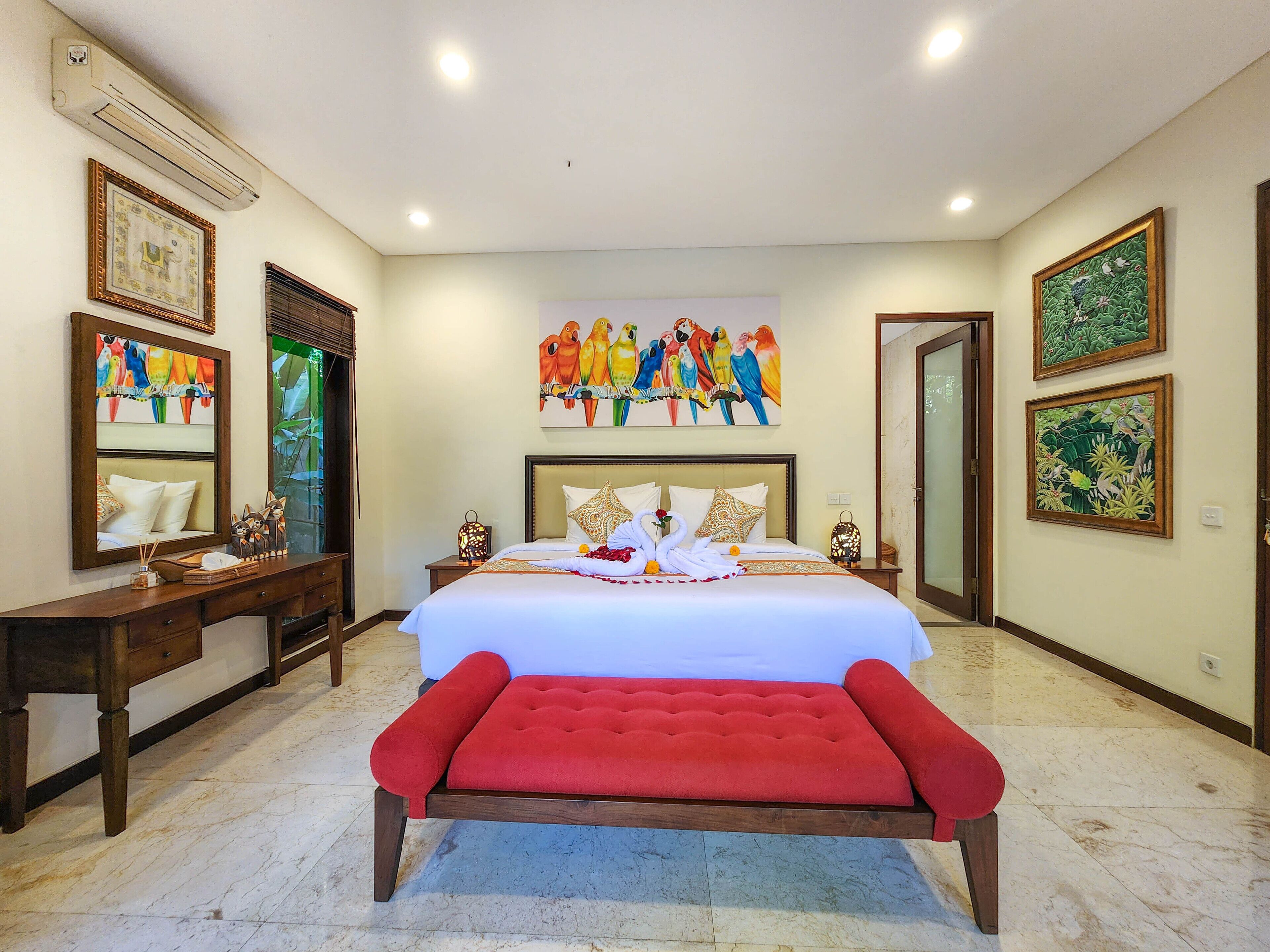 A hotel room with a large bed, a red bench, a wooden dresser, and artwork on the walls.