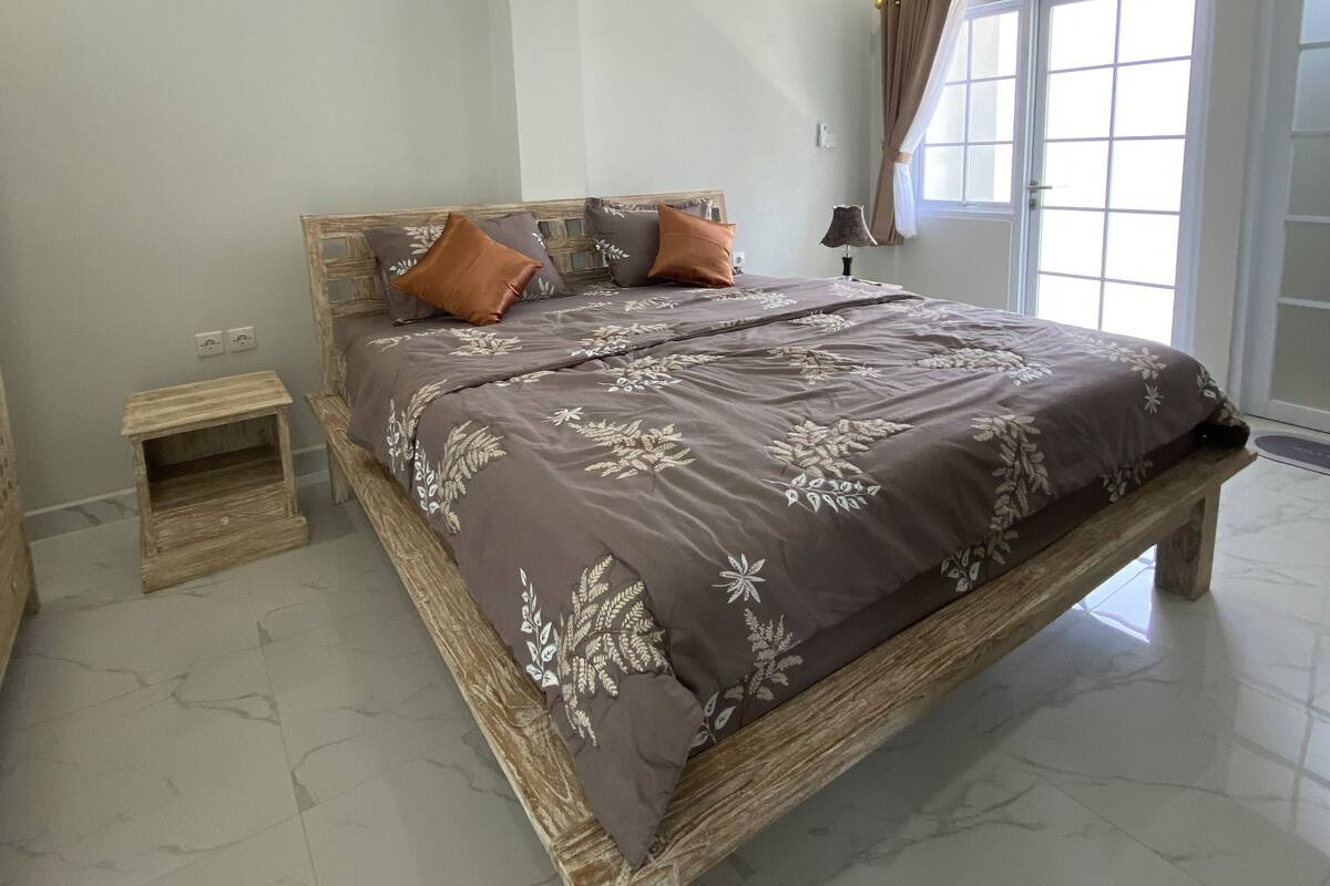 Ein Schlafzimmer mit einem hölzernen Bettgestell, einem kleinen Nachttisch und einem großen Fenster mit Vorhängen.