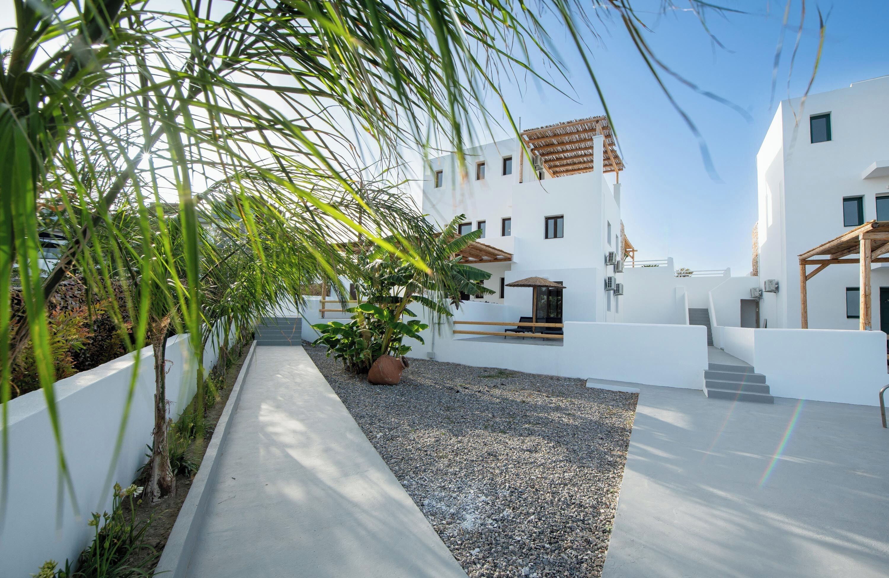 Un bâtiment moderne aux murs blancs et au toit recouvert de tuiles, entouré d’une cour en gravier et de verdure.