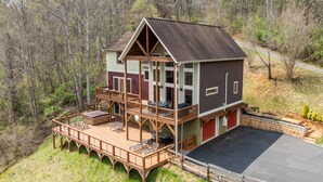 Une cabane à deux étages avec une terrasse et un garage, entourée d’une zone boisée.