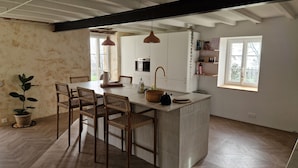 Uma cozinha com ilha central, cadeiras de madeira, luminárias pendentes e um vaso com planta.