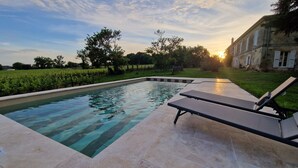 Piscina retangular com água cristalina, rodeada por um deck revestido com azulejos. Há duas espreguiçadeiras no deck. Ao fundo, uma área gramada com árvores e um edifício com telhado inclinado.