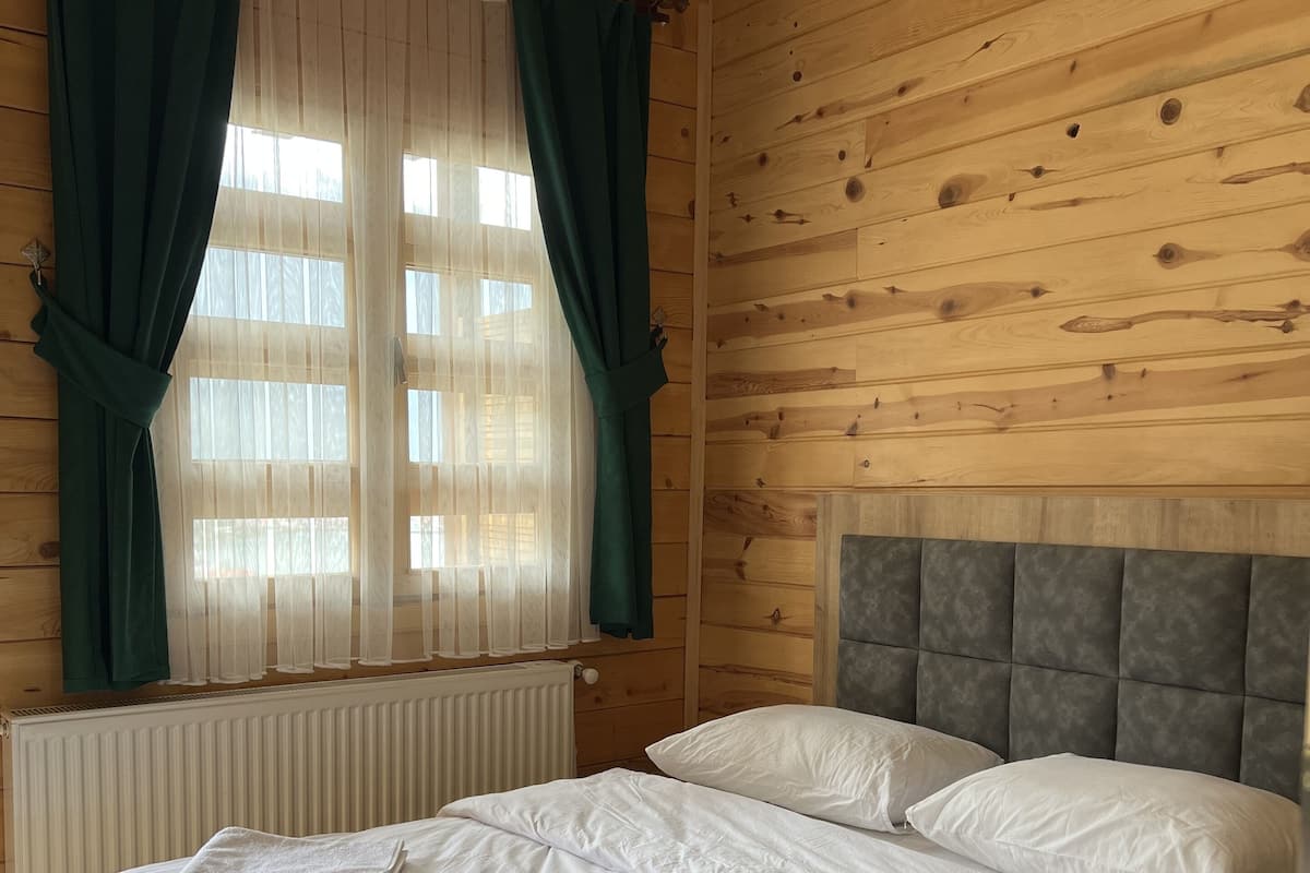 A wooden cabin room with a bed, green curtains, and a window with white curtains.