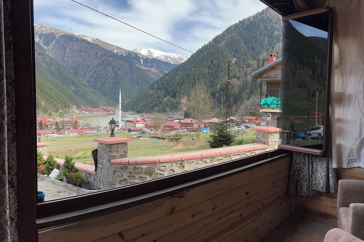 A room with a wooden interior, a large window offering a view of a mountainous landscape, a stone wall, and a small balcony with a red roof.