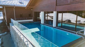 Piscina al aire libre con agua azul cristalina, rodeada por una terraza de madera y barandillas de vidrio.