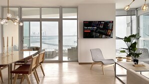 Una sala de estar moderna con un gran ventanal que da a un balcón, un comedor con muebles de madera y un televisor montado mostrando la interfaz de Netflix.