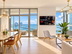 Un salon moderne doté d’un grand téléviseur, d’un coin repas avec une table en verre et offrant une vue sur les toits de la ville et sur l’eau.