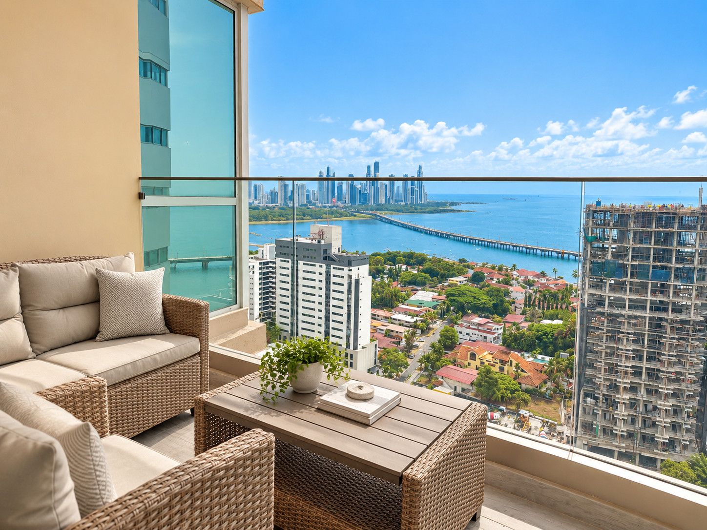 Ein Balkon mit einer Couch, Korbstühlen und einem Tisch mit einer Pflanze. Durch die Scheiben ist eine Stadtansicht und ein Blick auf das Wasser zu sehen.