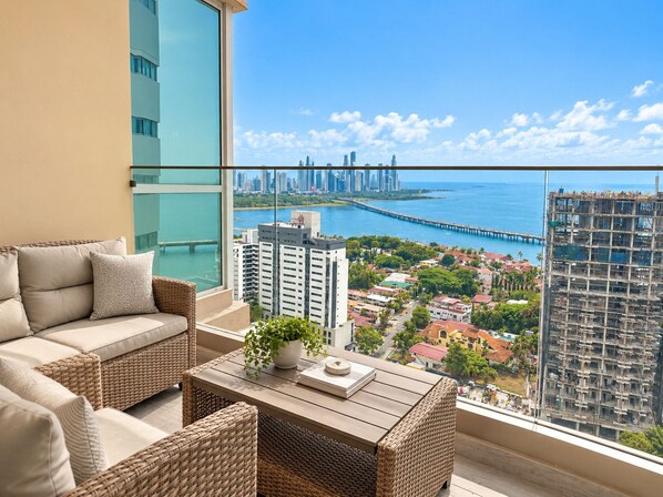 A balcony with a sofa, wicker chairs, and a table with a plant. A cityscape and waterfront are visible through the glass.