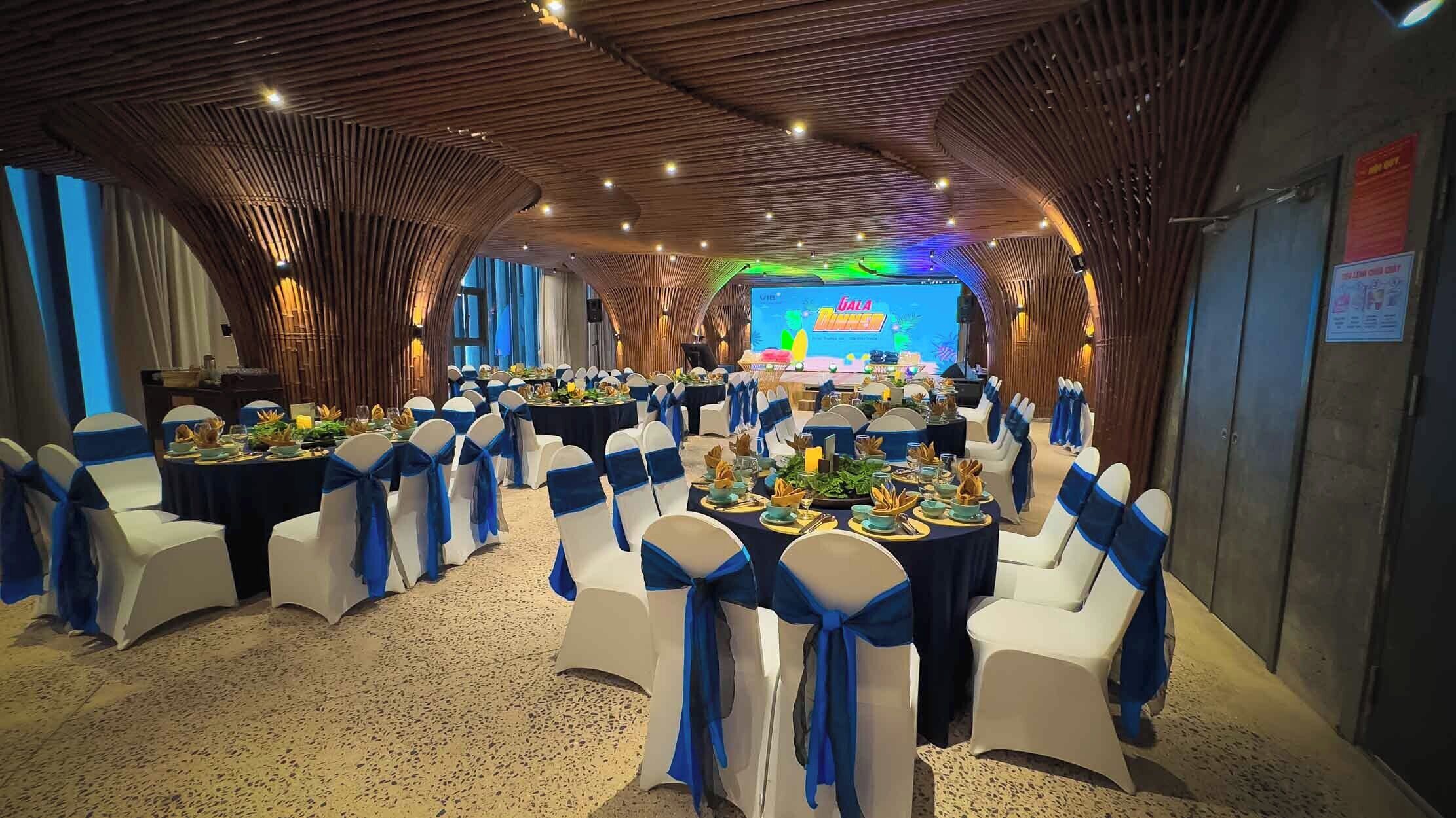 Une salle de banquet avec des tables rondes, des chaises blanches ornées de rubans bleus et une scène dotée d’un décor coloré.