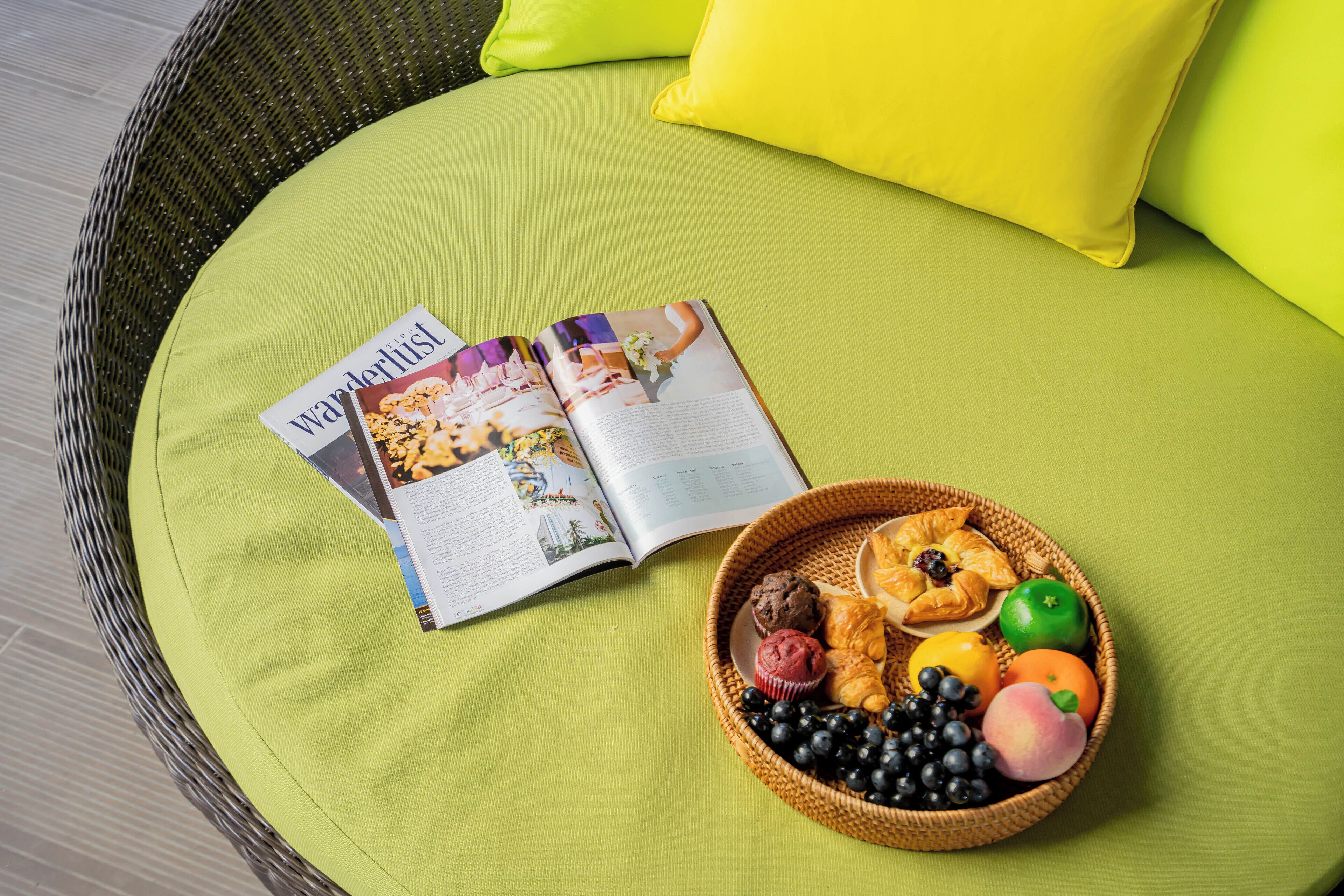 A wicker chair with a bright green cushion, a magazine titled "Wanderlust," and a basket of pastries and fruit.