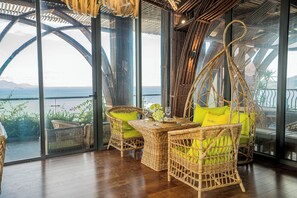 Een zithoek met rieten meubels, een tafel en stoelen, grote glazen deuren en uitzicht op zee.