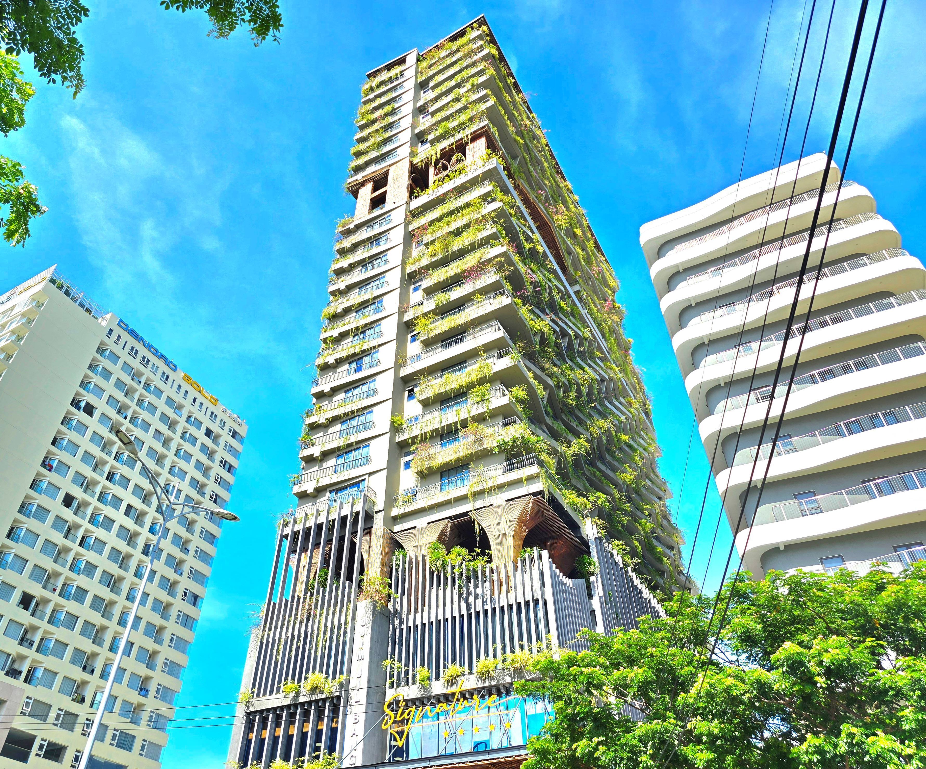 A high-rise building with multiple balconies, some with greenery, and a clear blue sky in the background.