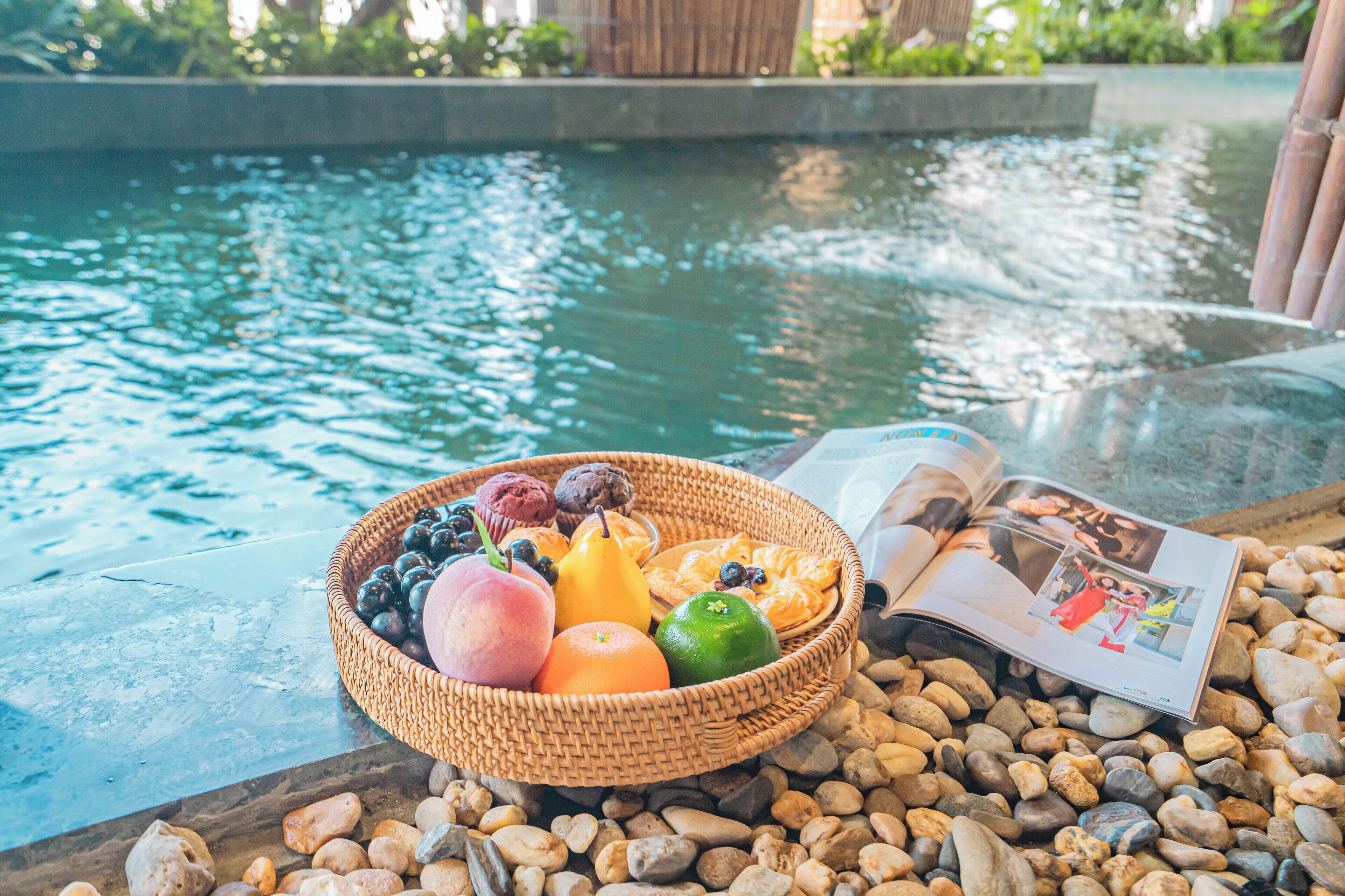 A wicker basket filled with fruits and pastries on a table by a pool, with a magazine open beside it.