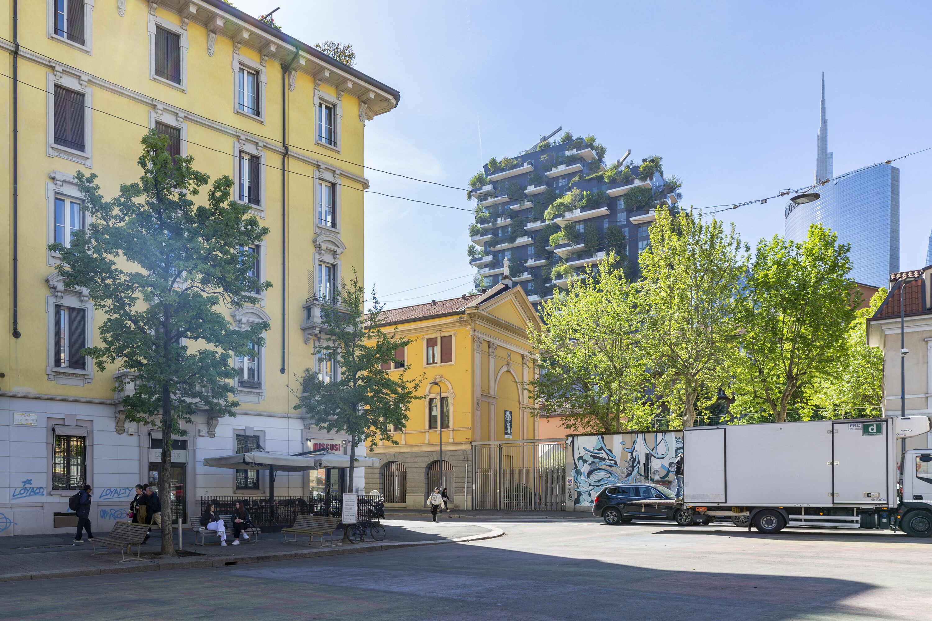 A street with a yellow building, a tree-lined sidewalk, and a modern high-rise in the background.