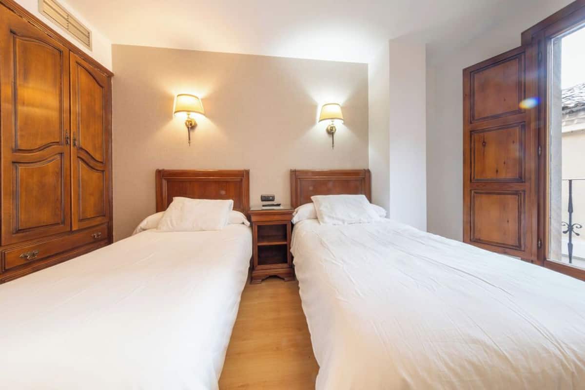A hotel room with two single beds, wooden wardrobes, and wall-mounted lamps.
