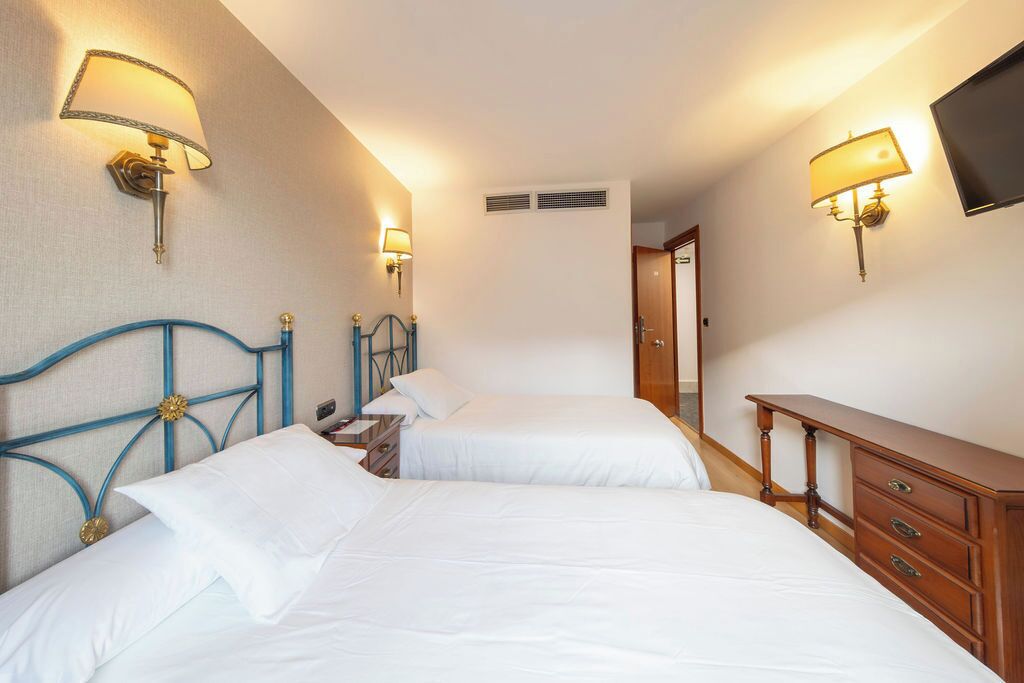 A hotel room with two beds, a wooden dresser, and wall-mounted lamps.