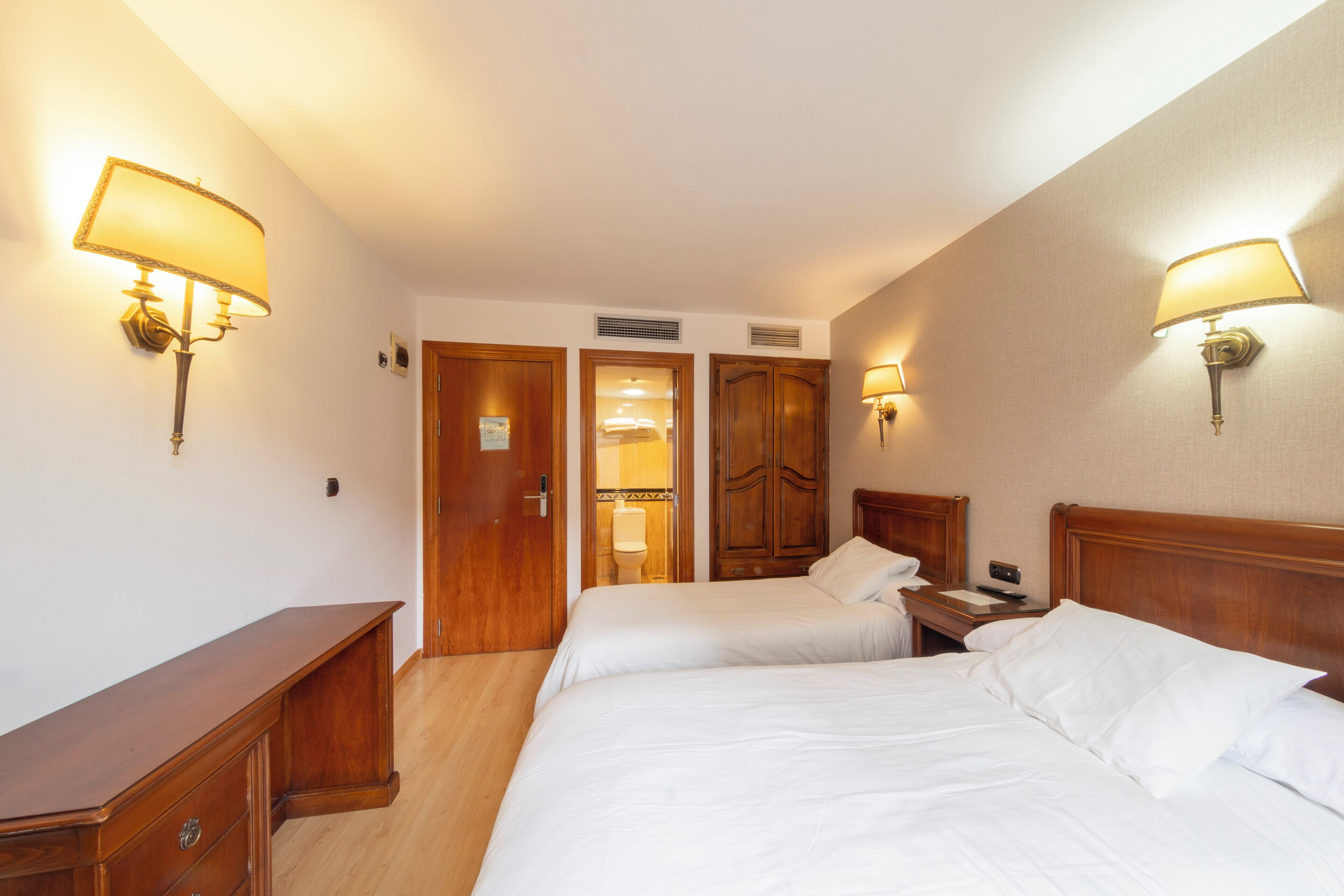 A hotel room with two beds, wooden headboards, and wall-mounted lamps.