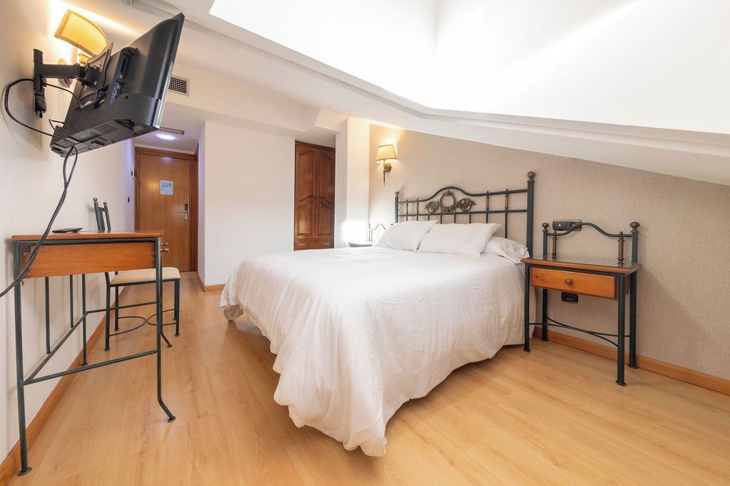 A bedroom with a bed, a desk, a TV mounted on the wall, and a door in the background.