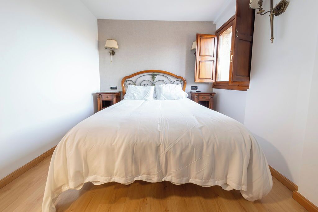 Een net opgemaakt bed met een houten hoofdbord, twee nachtkastjes en een wandlamp.