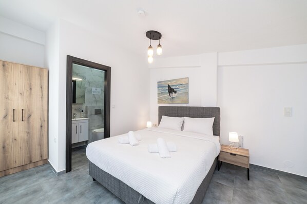 A modern bedroom with a large bed, a wooden wardrobe, a bathroom with a glass door, and a painting on the wall. - Olympiada Residential Complex & Villas (Pangaio)