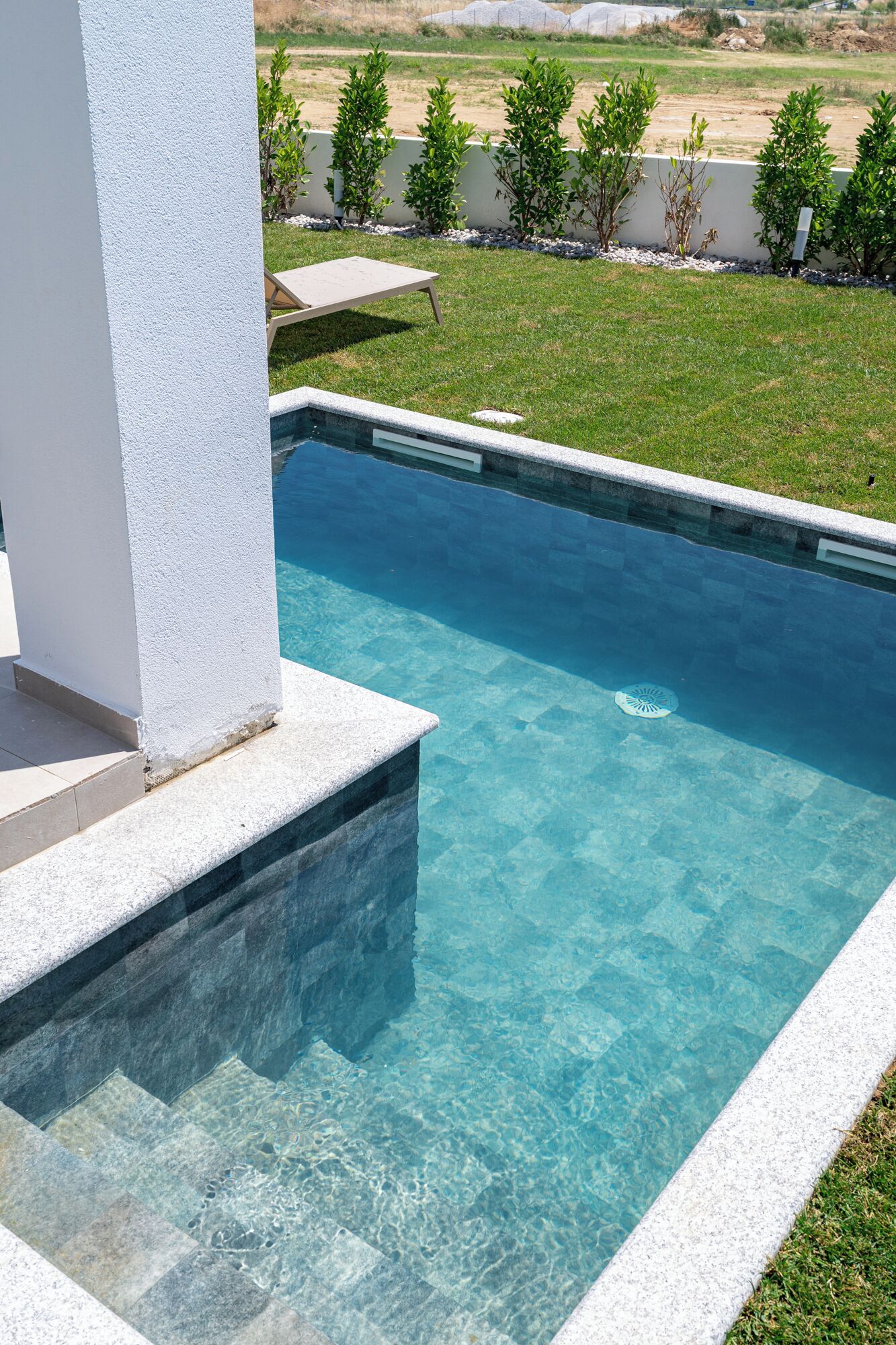 A rectangular swimming pool with clear blue water, surrounded by a white wall and a grassy area with small trees.