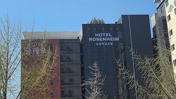 A modern building with 'HOTEL ROSENHEIM' signage, a parking area, and a street with a parked car and a motorcyclist.