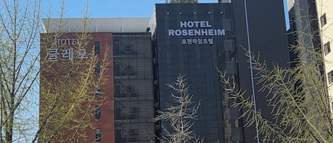 A modern building with 'HOTEL ROSENHEIM' signage, a parking area, and a street with a parked car and a motorcyclist.
