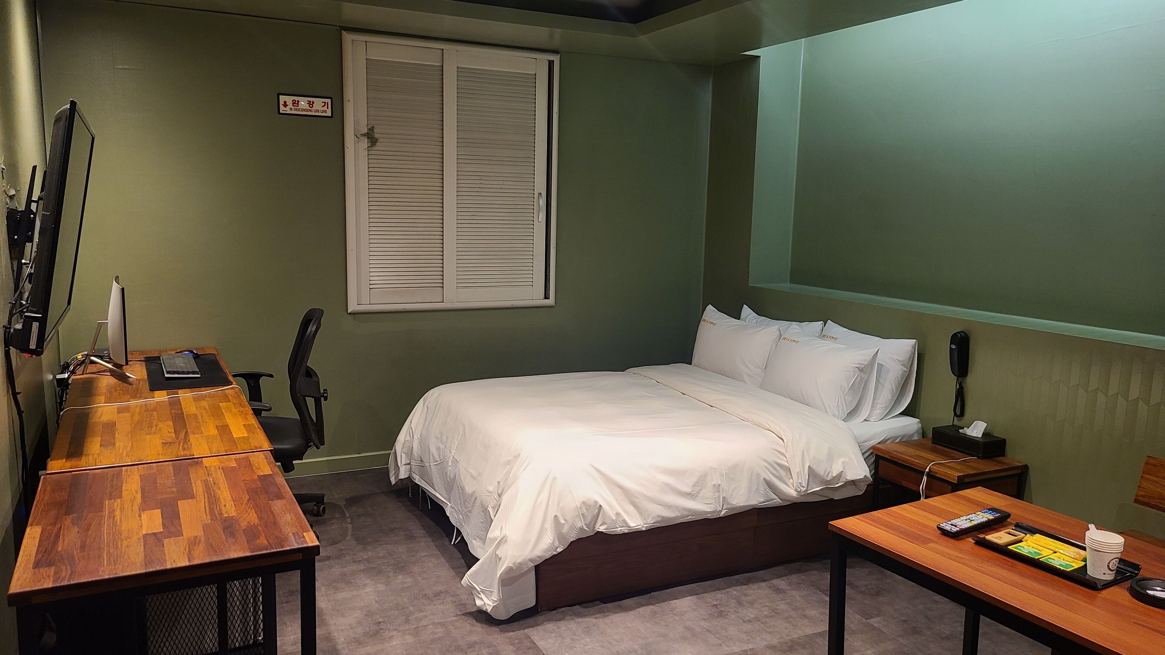 A bedroom with a bed, a desk, a chair, a bedside table, a telephone, and a window with blinds.