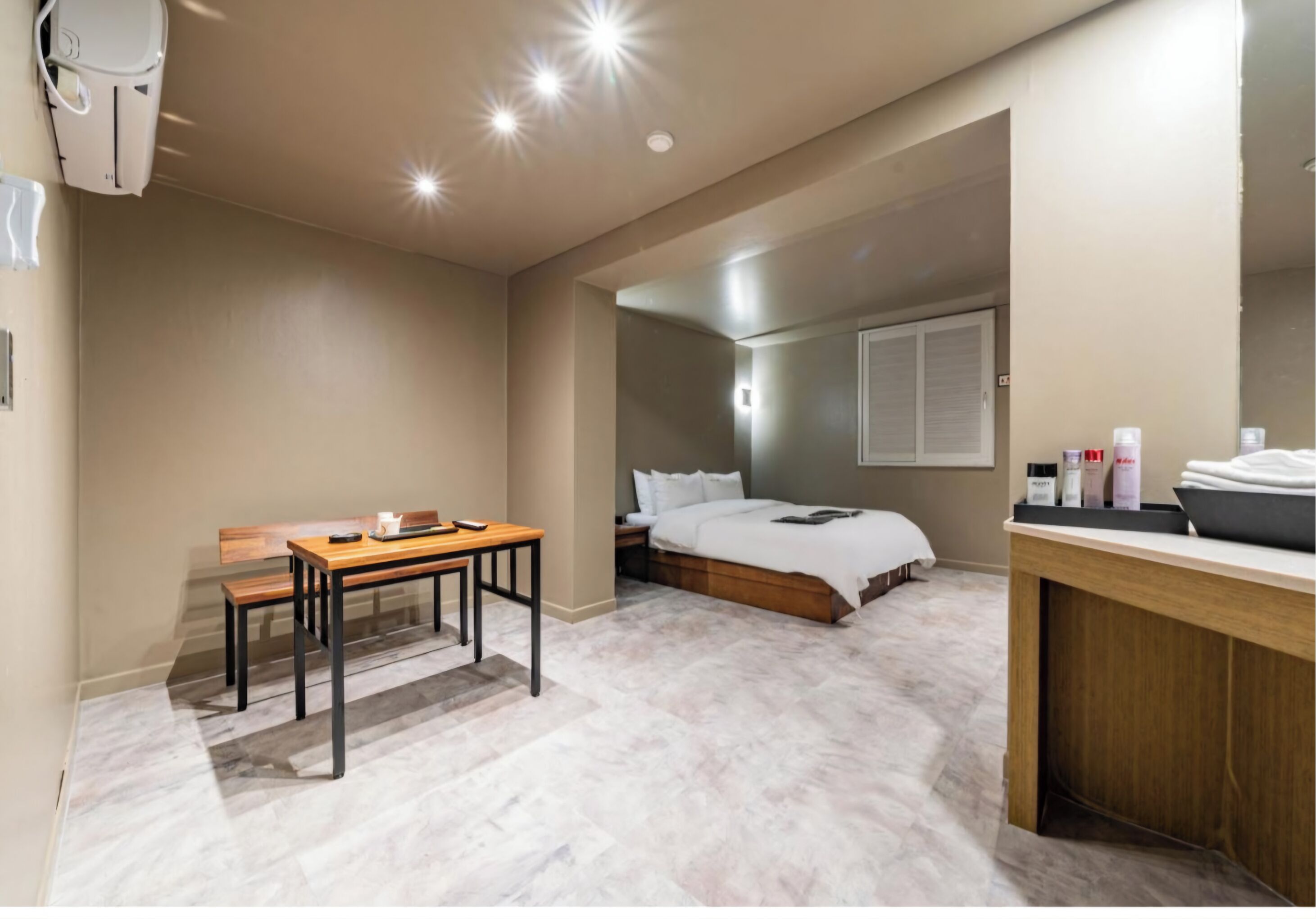A bedroom with a bed, bedside table, and a small desk with a chair. There is a bathroom with a sink and mirror.
