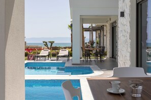 Zona de piscina con sillones de descanso y una mesa, con vista despejada al mar y una pared de piedra.